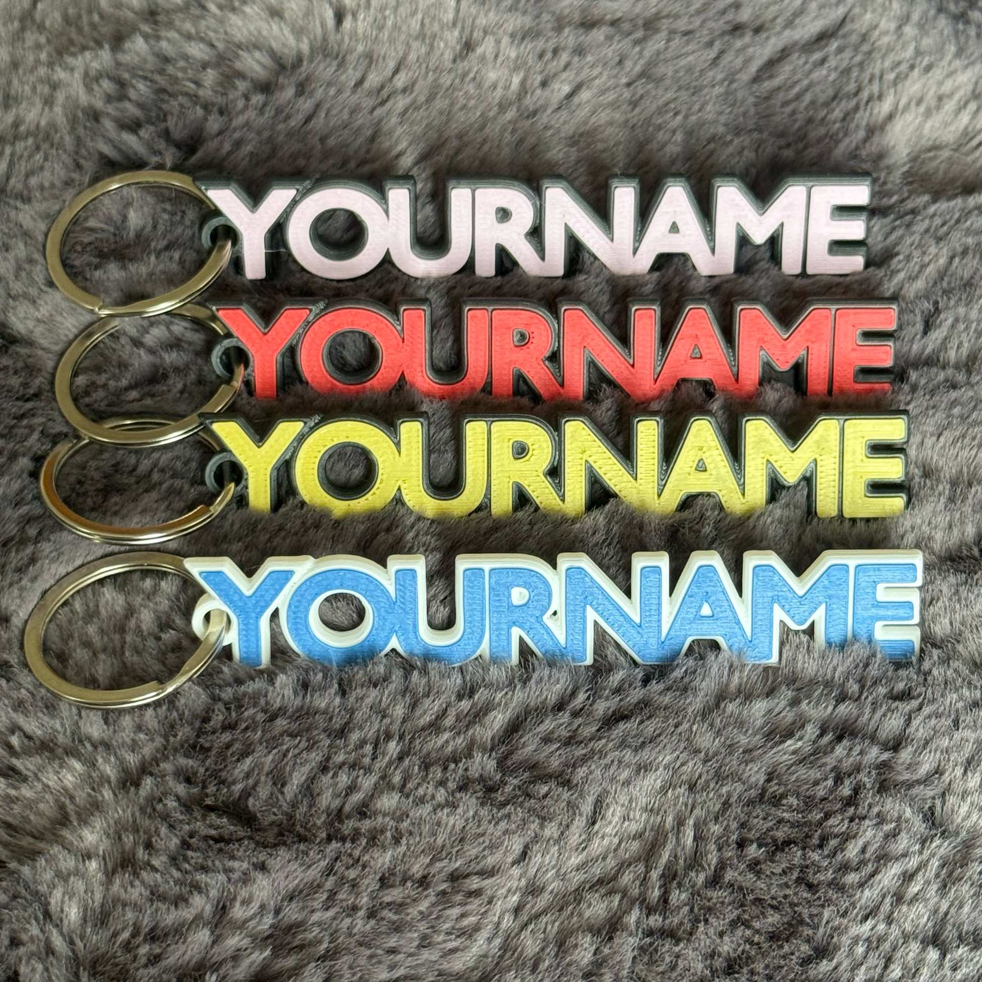 Four keychains with 'YOURNAME' text in different colors on a textured gray background