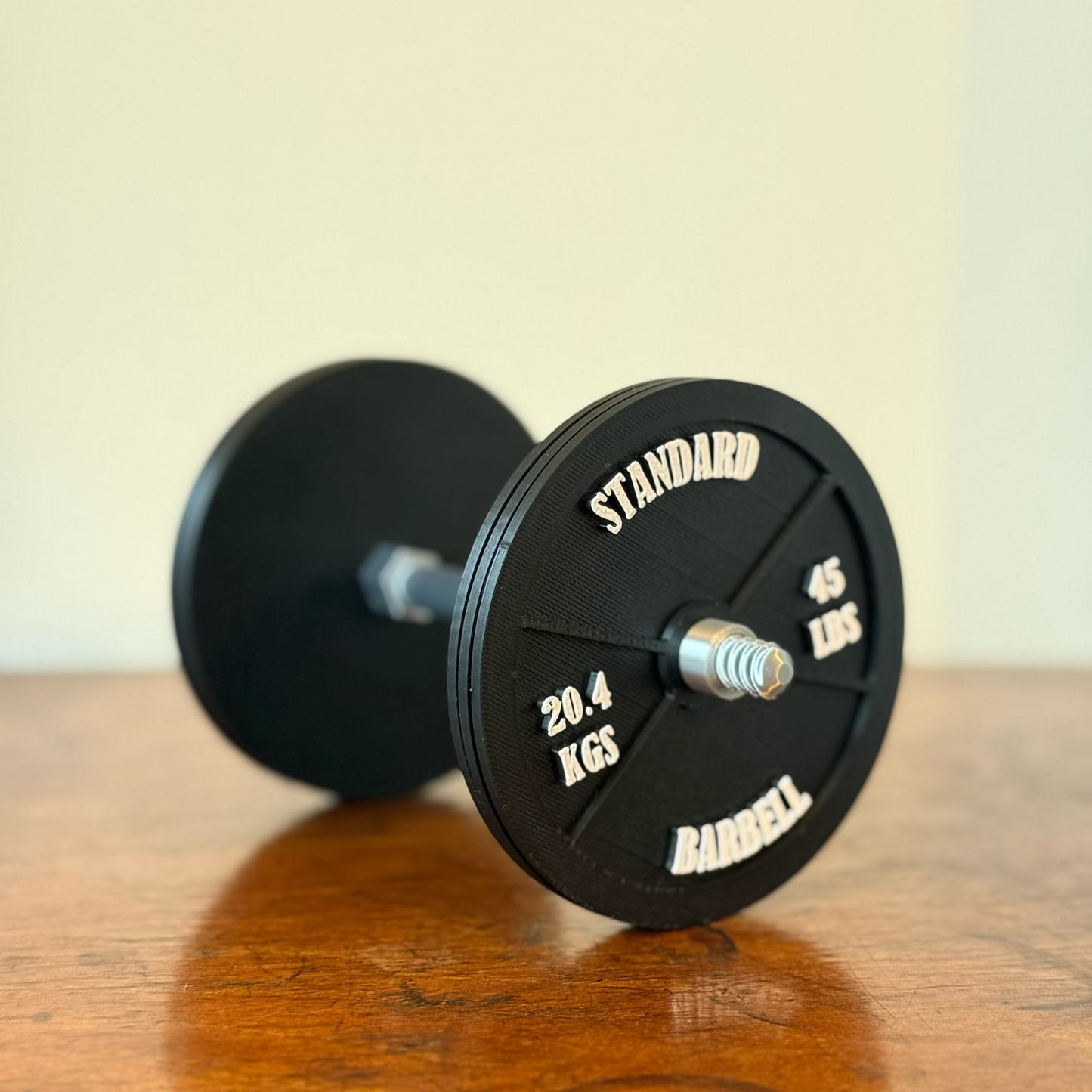 Realistic black dumbbell shaped coaster for gym lovers, 3D printed fitness-themed drink coaster, masculine home gym decor, weight plate desk accessory for bodybuilders placed on a wooden table.