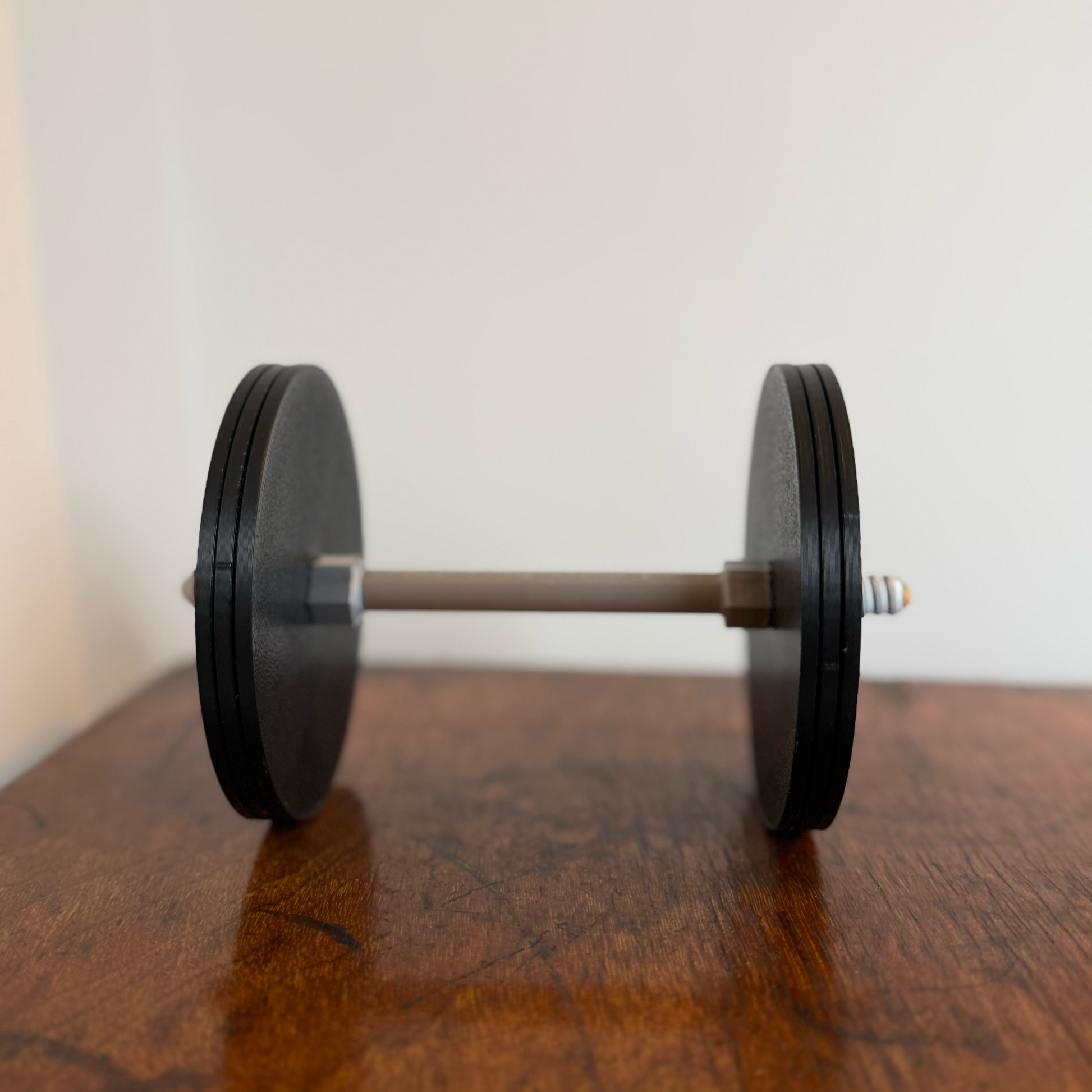 Realistic black dumbbell shaped coaster for gym lovers, 3D printed fitness-themed drink coaster, masculine home gym decor, weight plate desk accessory for bodybuilders placed on a wooden table.
