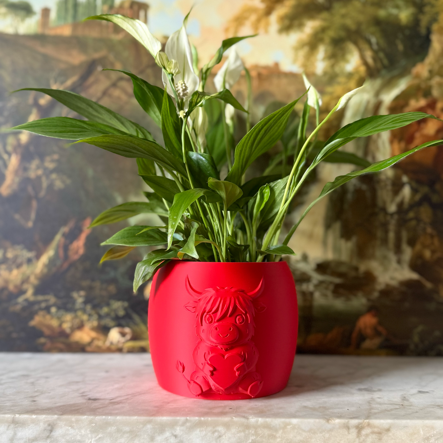 Red highland cow valentines plant pot with the cow holding a heart in its arms against a painting background on a marble top surface