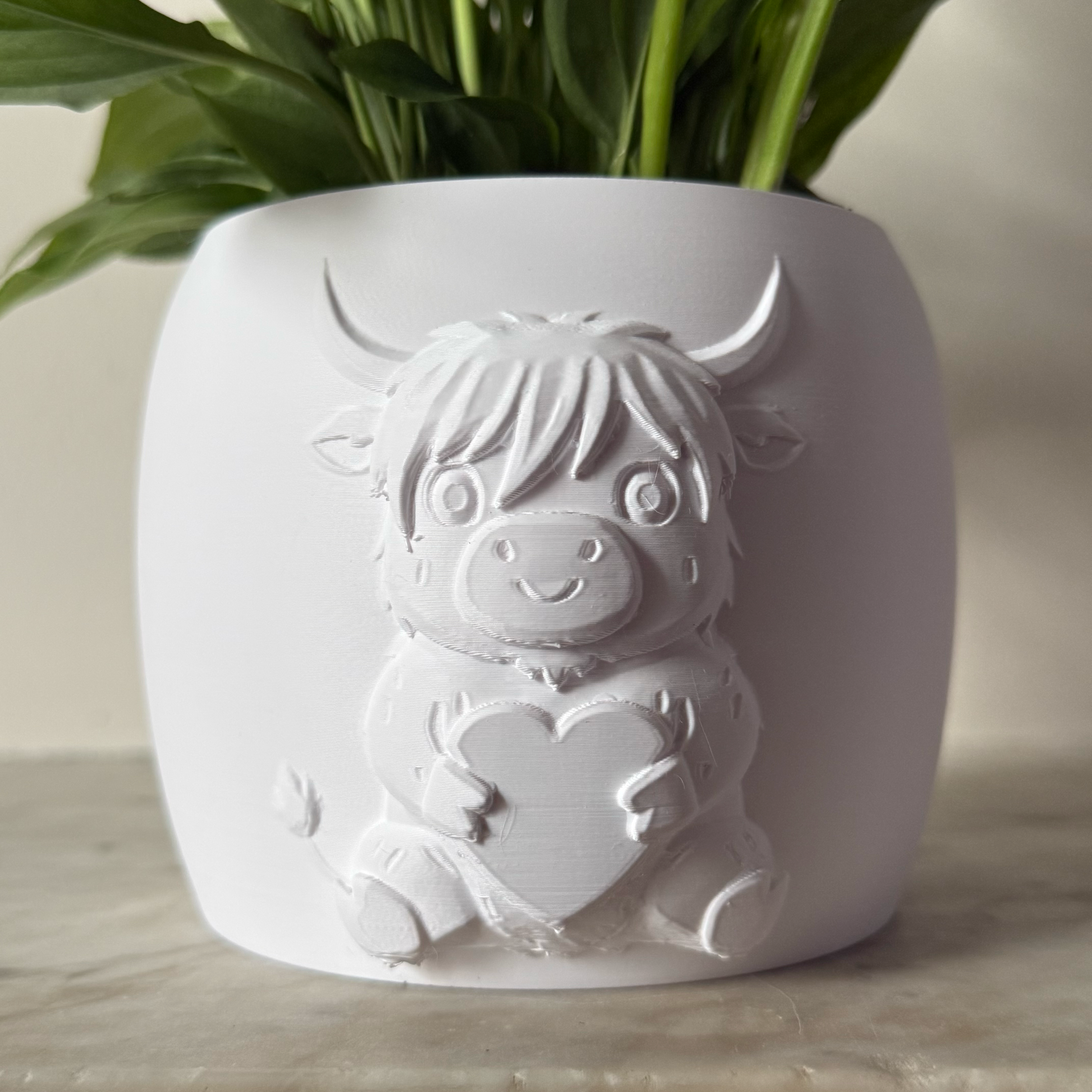 Potted plant in a decorative white highland cow pot on a surface with a scenic painting in the background