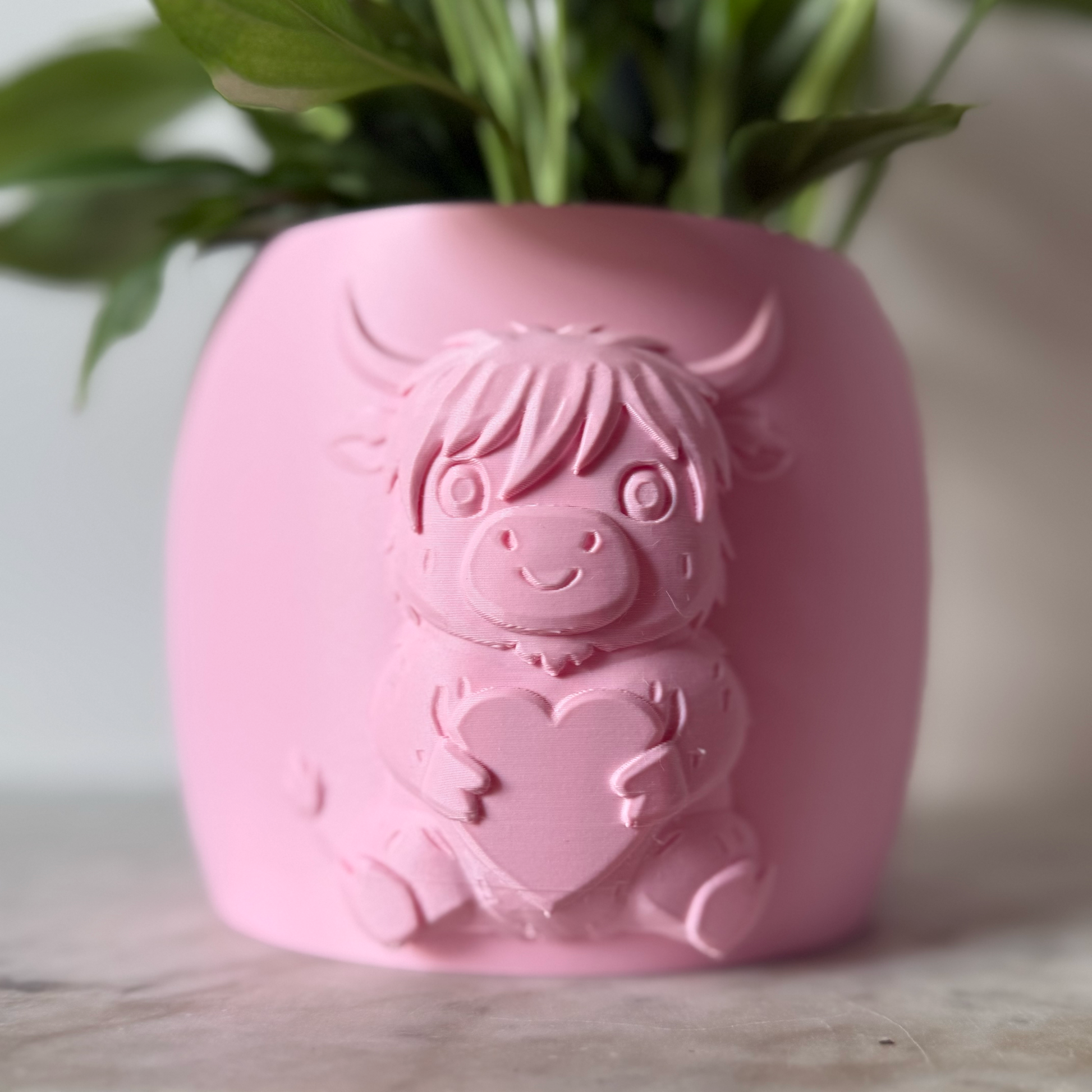 Pink highland cow valentines plant pot with the cow holding a heart in its arms against a white background on a marble top surface
