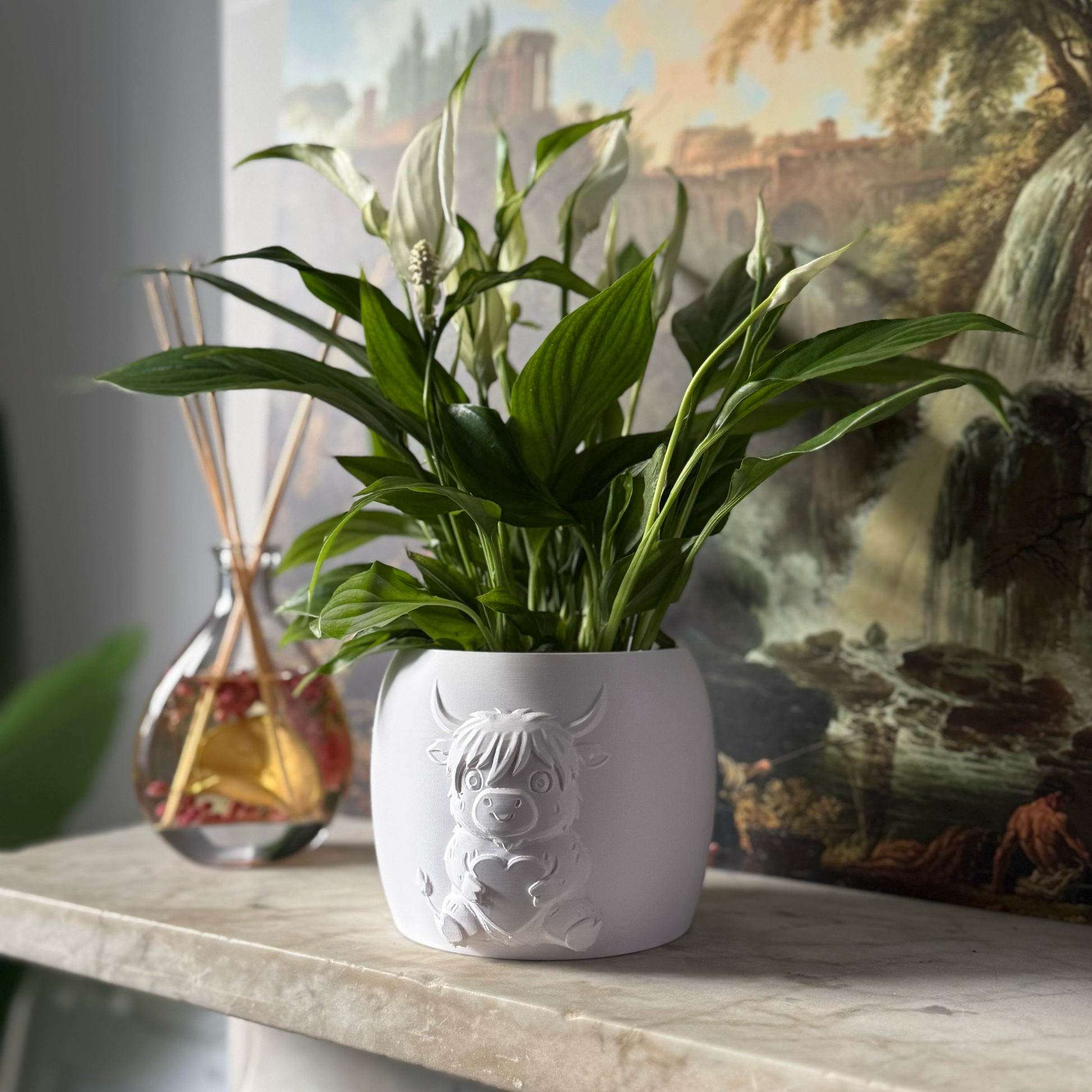 Potted plant in a decorative white highland cow pot on a surface with a scenic painting in the background