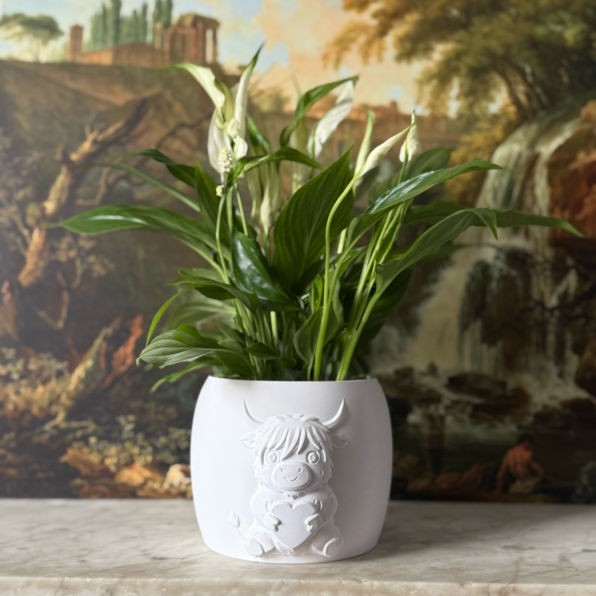 Potted plant in a decorative white highland cow pot on a surface with a scenic painting in the background