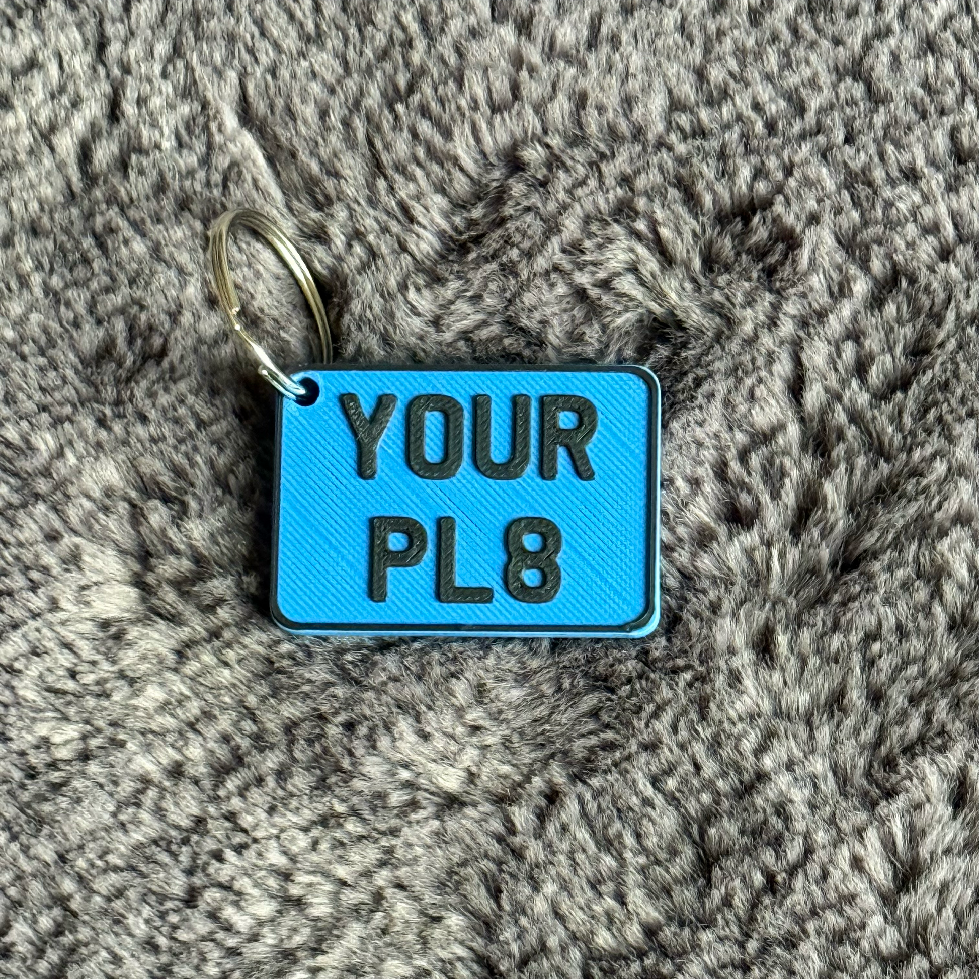 Blue keychain tag with 'YOUR PL8' text on a textured gray surface