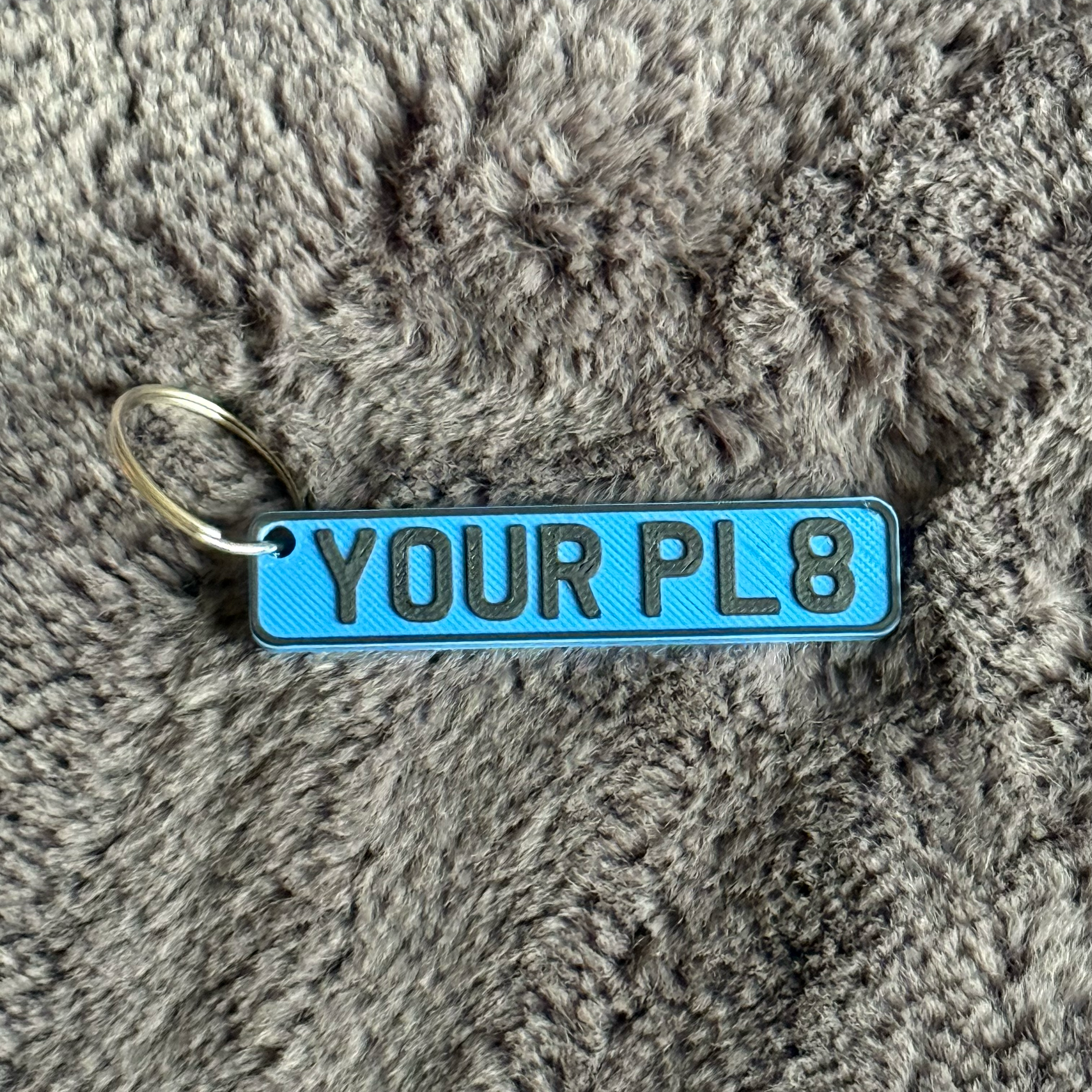 Blue keychain with 'YOUR PL8' text on a textured gray surface