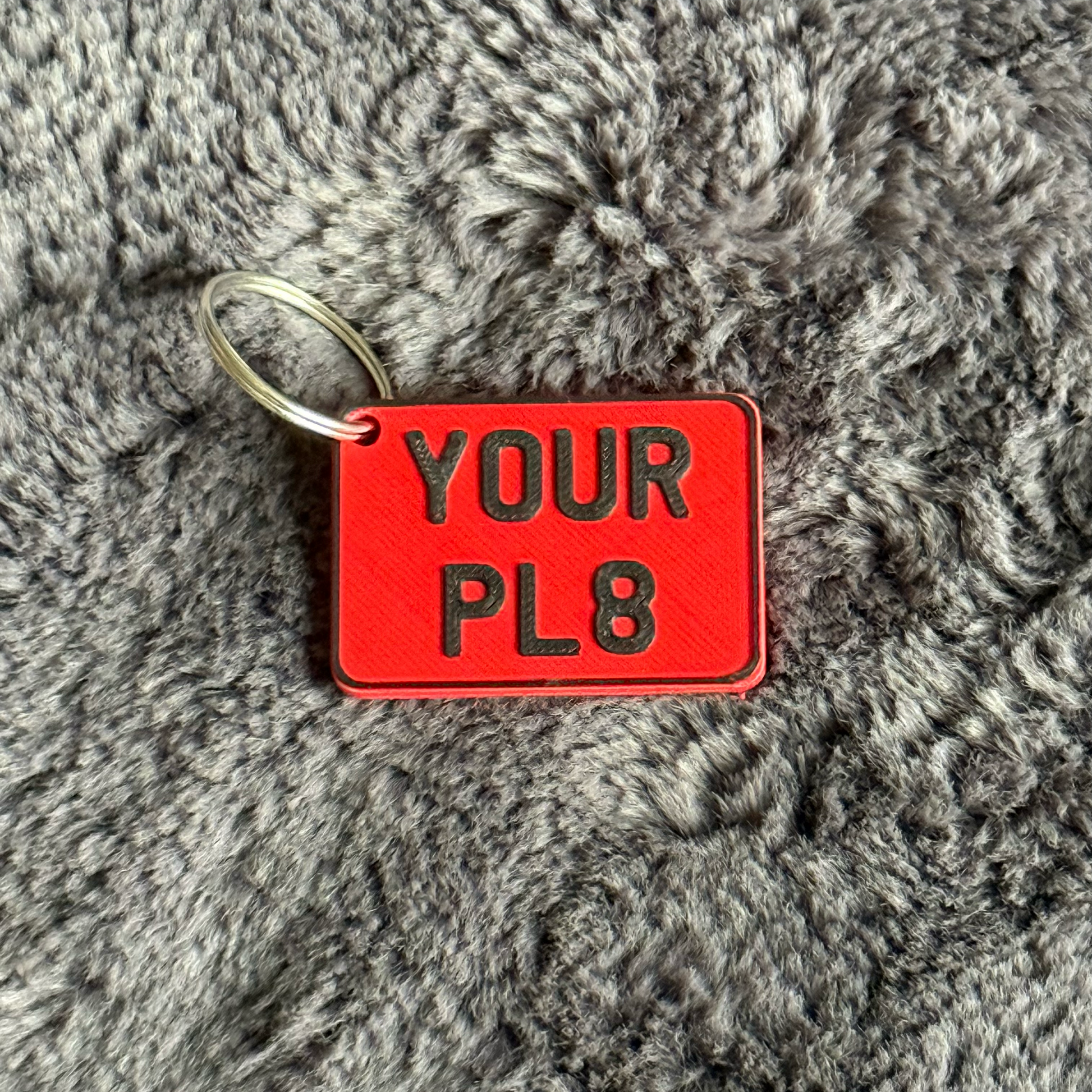 Red rectangular keychain tag with 'YOUR PL8' text on a gray textured background