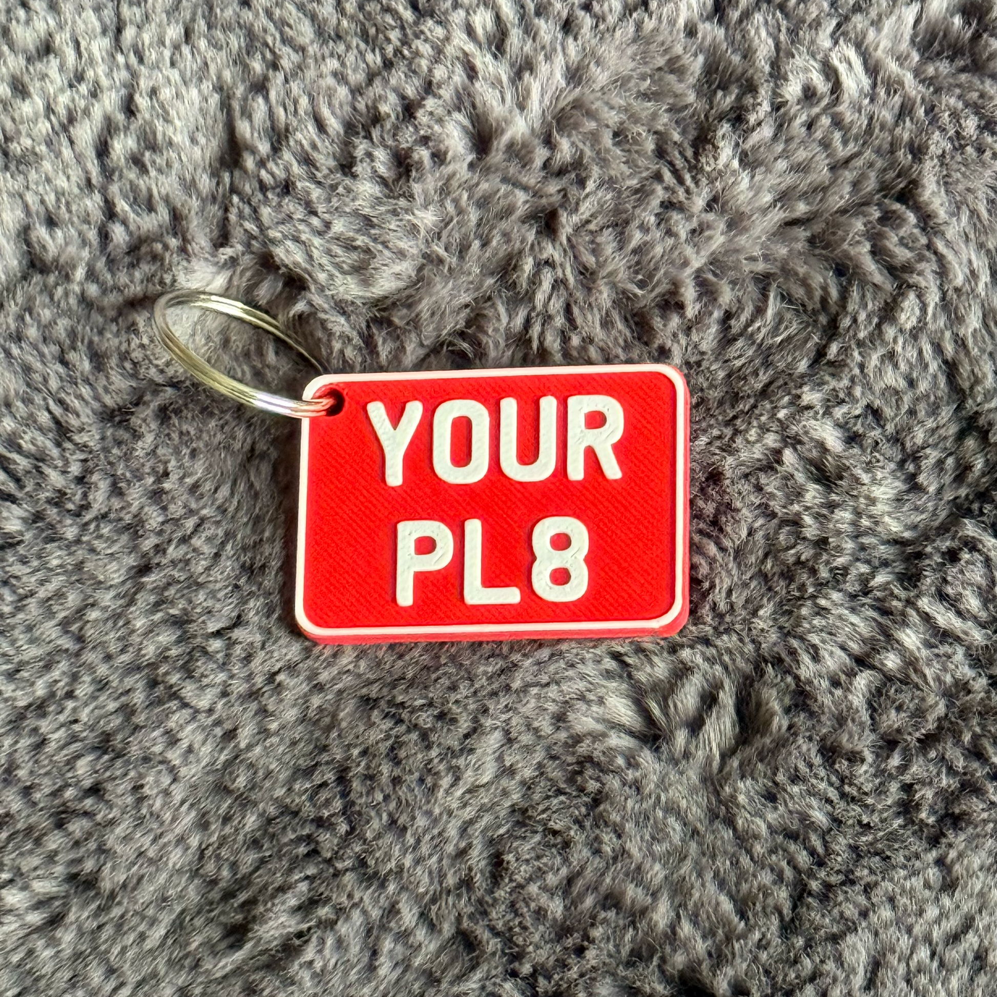 Red keychain tag with 'YOUR PL8' text on a gray textured background